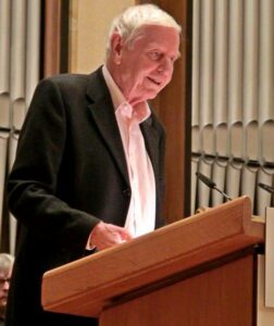 Side profile of Hans Magnus Enzenberger in a black and white suit. He is speaking on lectern with organ pipes behind him.
