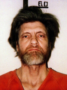A color forward facing Kaczynski’s mugshot. He has a light mixed gray and black beard. He is wearing an orange jumpsuit. His black medium guy length hair just touching 60 inches.