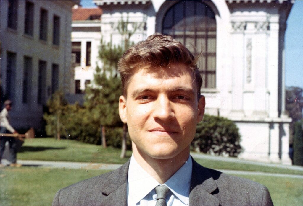 A color landscape photograph of Ted Kaczynski as a student with a façade of a white building behind him. 