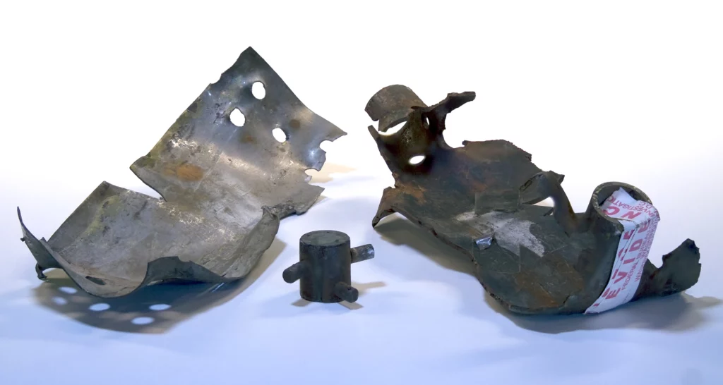 A color photograph of three pieces of shrapnel from one of Kaczynski’s bombs. All of the pieces are placed concave up on a blue background