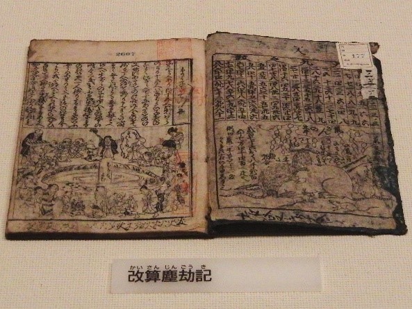 A photograph of open book with kanji. The pages are weathered. There is a drawing on the bottom right of the page of a pond surrounding by people