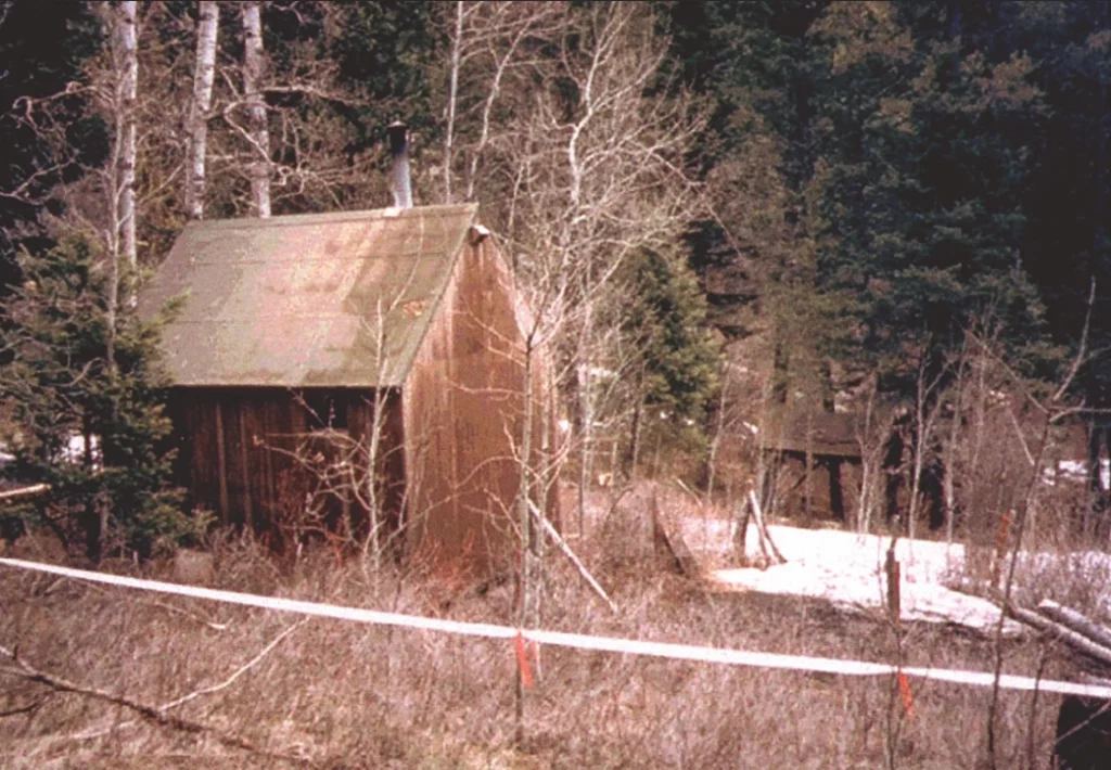 A color landscape photograph of the cabin near Lincoln, Montana. The cabin is off the left with an open space filled with some snow to the right of it. The background is a dark green forest.