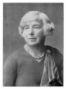 A black and white film portrait of Margaret Murray taken in 1934. She has mid-length hair and is wearing a necklace, a sash and a dress. She is facing to the left.