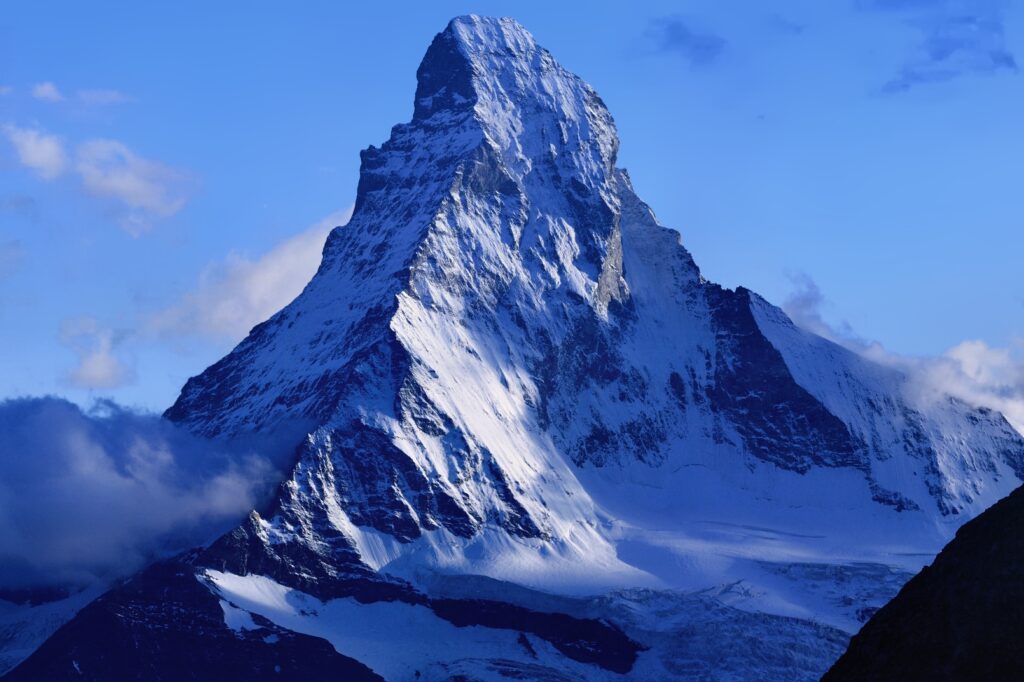 A photograph of the Matterhorn in Switzerland. It is shaped like a pointy hat and is covered in snow. Some clouds a visible lower down the mountain. 