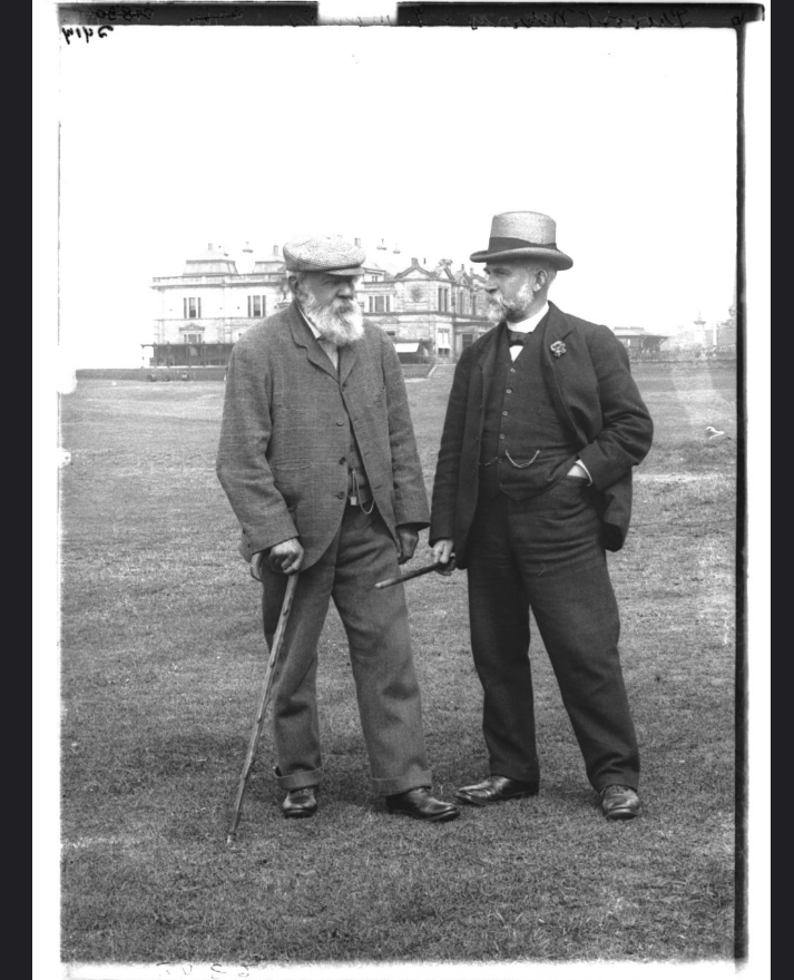 A photograph of Old Tom Morris and Provost George Murray in 1905. Both are dressed in suits and have beards. Old Tom Morris is holding a walking stick for support, and they are both standing on the 18th fairway of the Old Course with the R&A Clubhouse visible in the background. 