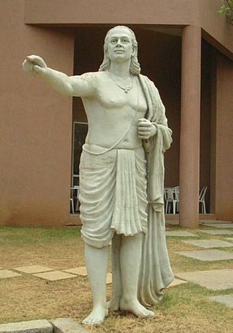 A color photograph of marble Statue of Aryabhata reaching their right hand pointing. The statue is wearing robe covering the waist below and the left shoulder. The statue is outside in the garden. 