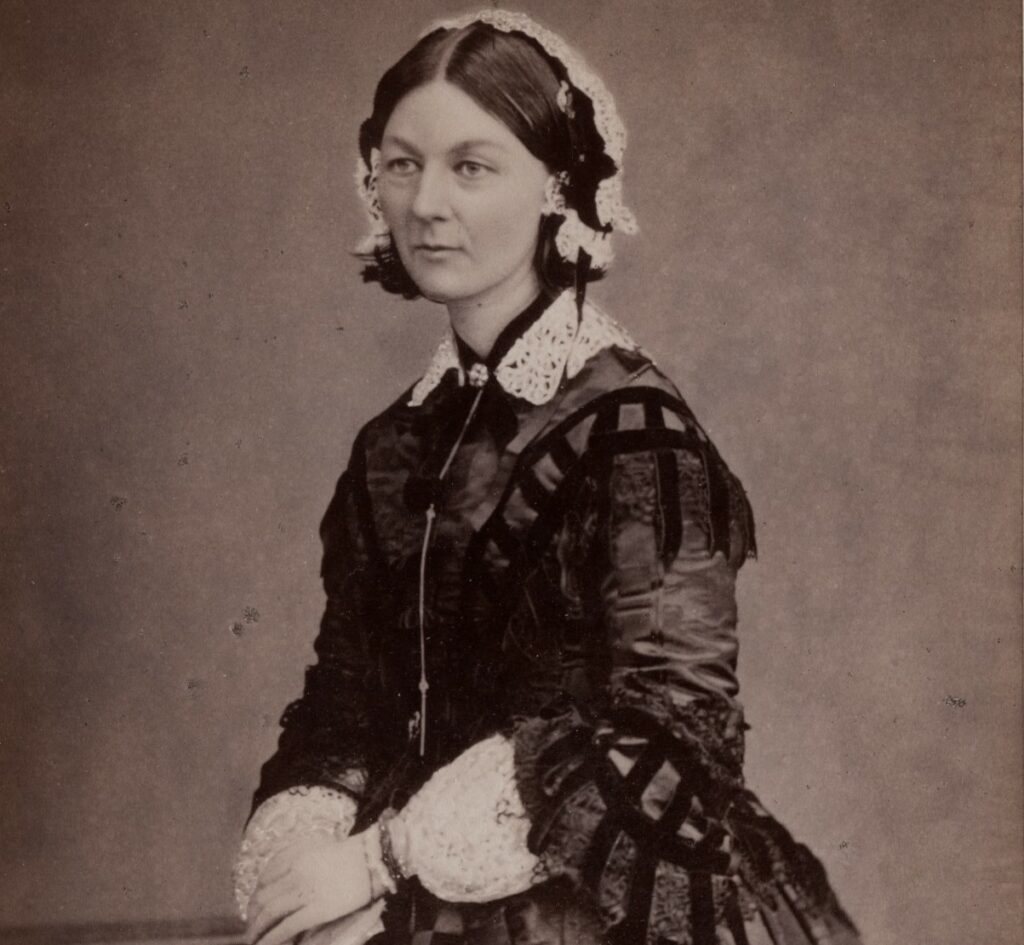 A black and white portrait of Florence Nightingale standing in front of a plain background. She has her hands clasped together and is wearing a Victorian dress. The photograph is taken above the waist while Florence is sitting on a stool.