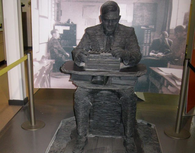 An image showing a black stone statue of Alan Turing working with an enigma machine. 