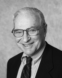 A black and white portrait showing Kenneth Arrow. He is wearing a suit and tie, glasses, and is smiling. 
