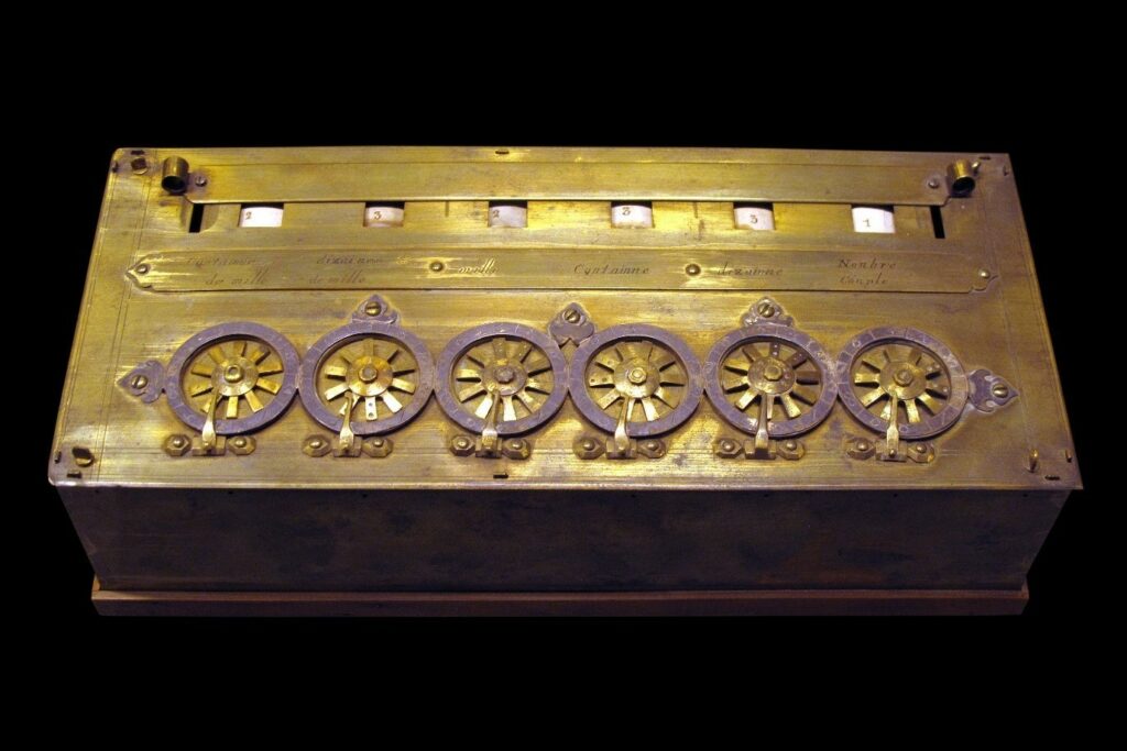 An photograph of a surviving example of a Pascaline. It is rectangular and gold coloured, although in need of a polish. It consists of six number displays parallel to its top edge and six round dials parallel to its bottom edge. 