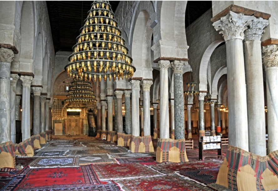 An image showing white stone arches held up by elaborately decorated stone columns within the Great Mosque of Kairouan. Between the stone columns hang two chandeliers, while a patchwork of carpets form the floor of the mosque. 