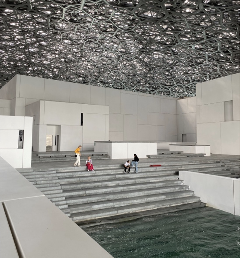 An image showing an open space within the Abu Dhabi Louvre. Stone steps lead down to a small pool, while people are sitting on the steps. Above them is an elaborate roof which looks like a birds nest. 