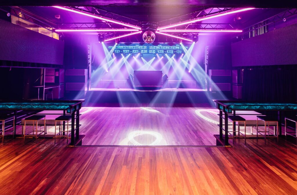 A photograph of a dancefloor. There is  a raised bar area with steps leading down to the dancefloor. A stage is located behind the dancefloor, with DJ equipment sitting on it. Strobe and LED lights are suspended from the ceiling and are illuminating the dancefloor. 