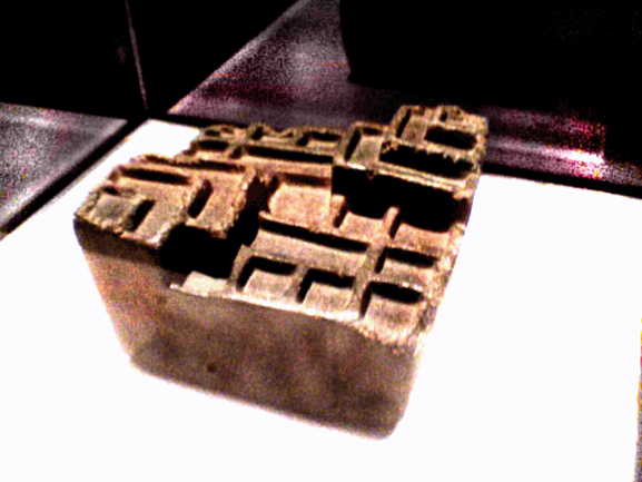 Image shows an object in a glass case, on a white platform. It is brown and shaped like a cuboid with other square blocks and ridges on the top, in a geometric pattern.